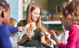 A woman smiles while holding clay, surrounded by children crafting with colorful modeling clay in a classroom setting.