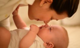 An adult gently leans closer to an infant, their noses almost touching. The baby gazes upward, lying on a soft surface, creating an intimate, playful atmosphere.