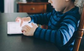 A child, wearing a blue striped shirt, touches a tablet screen with both hands, seated at a dark wooden table in a dimly lit room.