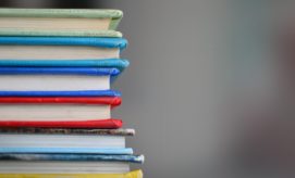 A stack of seven colorful books sits on a flat surface, with a blurred gray background providing a neutral setting.