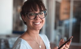 A person with glasses smiling while holding a smartphone, standing in a softly lit indoor space with blurred background details.