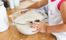 A child mixes batter in a glass bowl using a whisk, sitting on a wooden table surrounded by kitchen items in a warmly lit room.