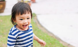 A smiling child, wearing a blue and white striped shirt, stands on a grassy area with concrete paths in the background, appearing joyful and expressive.