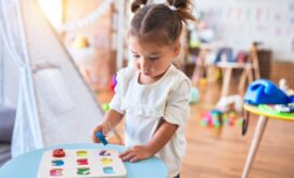 A child plays with a colorful alphabet puzzle on a blue table, focusing intently. Nearby, a playroom filled with toys and a teepee tent creates a cheerful, nurturing environment.