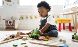 A toddler, sitting on a carpet, plays with a green toy train on a wooden track in a brightly lit playroom, surrounded by various toys and a colorful bag.