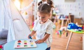 A young child with brown pigtails places a blue letter into a puzzle on a light blue table in a brightly lit playroom filled with toys and drawings.