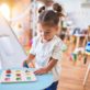 A young child with brown pigtails places a blue letter into a puzzle on a light blue table in a brightly lit playroom filled with toys and drawings.