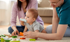 A baby, chewing a toy block, is surrounded by colorful blocks on a carpeted floor, while two adults attentively interact with the child, suggesting a playful family setting.