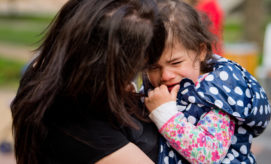 Child crying in a flowered outfit and polka-dotted jacket, being comforted by a person with long hair, outdoors with blurred greenery in the background.