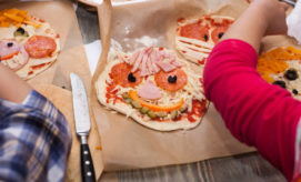Children decorate small pizzas with various toppings like pepperoni, ham, cheese, and olives, creating smiley faces on dough. They work on parchment paper in a casual kitchen setting.