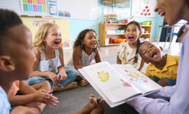 Female preschool Teacher Reads Story To Multi-Cultural Class Seated In Classroom