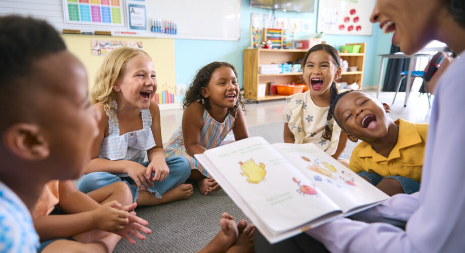Female preschool Teacher Reads Story To Multi-Cultural Class Seated In Classroom
