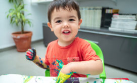 A young child, smiling and covered in colorful paint, engages in finger painting at a table. The setting is a bright room with a potted plant and bookshelves in the background.