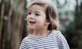 A young child with long brown hair smiles, wearing a striped shirt with dots, standing outdoors with blurred greenery in the background.