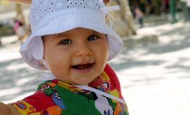 A smiling baby wearing a white sun hat and colorful shirt, reaching forward playfully, outdoors on a sunny day with blurred trees and people in the background.