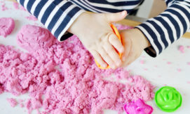 A child presses orange mold into pink kinetic sand on a white table, surrounded by various colorful molds. The child wears a long-sleeve striped shirt.
