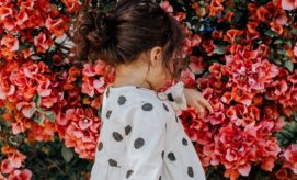 A young girl with brown hair in a bun, wearing a white dress with black polka dots, gazes downward, standing in front of a vibrant bush of pink flowers.