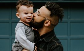 Adult kisses a smiling child on the cheek, holding them in a loving embrace. They are standing in front of a blue garage door with square panels, creating a warm, familial scene.