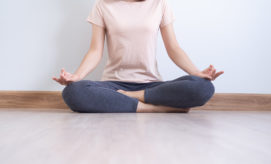 A person sits cross-legged on a wooden floor, meditating with hands in a mudra pose, wearing a light pink shirt and dark pants, against a plain light-colored wall.