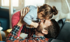 A person holds a child playfully at face level, both smiling, in a cozy living room with soft lighting and blurred furniture in the background.