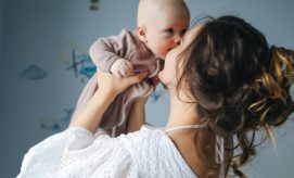 A woman holds a baby close to her face, gazing affectionately. The baby, dressed in a onesie, reaches towards her. They are in a softly lit room with wall decorations.