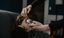 An adult hand uses a digital ear thermometer to check a child's temperature on a couch in a dimly lit room.