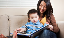 A young child, sitting on a woman's lap, looks at a book they're holding together on a beige sofa. The woman smiles, engaging with the child in a cozy room.