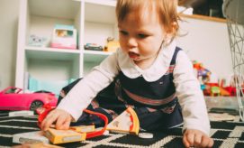 A toddler plays with toy food pieces on a patterned rug, surrounded by a pink toy car and shelves filled with books and colorful objects in a playroom.