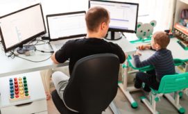 A man works at a desk with three monitors, while a child sits beside him at a small desk. Toys and office supplies clutter the brightly lit room.