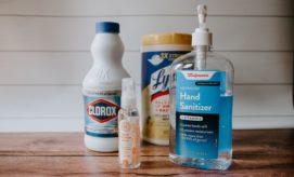 Bottles of disinfectant products, including Clorox, Walgreens Hand Sanitizer, and Lysol, are grouped on a wooden surface with a white paneled wall backdrop. Text on the sanitizer: "Walgreens Advanced Hand Sanitizer Vitamin E."