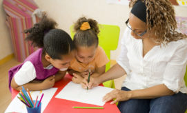 A woman helps two children draw on paper at a small red table. Colored pencils are in a cup nearby, and a colorful classroom environment surrounds them.
