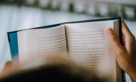 A person holds an open book, reading with focus. The surrounding environment appears softly lit, suggesting an indoor setting. Text on the visible page is partially blurred and unreadable.