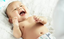 A baby smiles widely while lying on a soft, white blanket, wearing a patterned diaper. The surrounding area is softly lit, conveying a cozy and playful atmosphere.