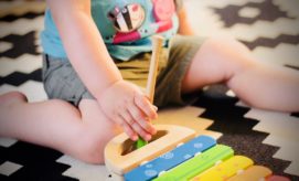 A child's hand plays with a colorful xylophone using a mallet, while seated on a patterned black-and-white rug, wearing shorts and a blue T-shirt with cartoon characters.