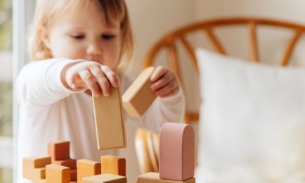 A child stacks wooden blocks on a table, focusing intently. The scene is set in a cozy room with a wooden chair and soft lighting in the background.