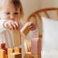 A child stacks wooden blocks on a table, focusing intently. The scene is set in a cozy room with a wooden chair and soft lighting in the background.