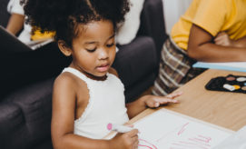 Child draws with a pink marker on white paper, sitting at a wooden table. A sofa and another child in a yellow shirt are in the background.