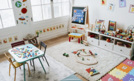 Children's playroom with a colorful table displaying toys, surrounded by chairs. A chalkboard titled "THE PLANET" stands nearby. Shelves hold educational toys, and a rug features a wooden train set.