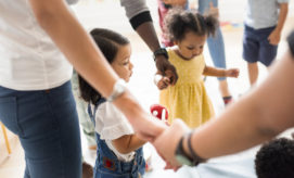 Children and adults hold hands in a circle, participating in an activity. The setting appears to be a bright, cheerful classroom with multiple people engaged in the background.