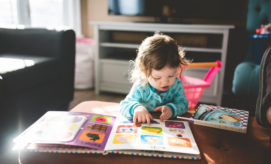 A child, seated at a table, points at a colorful open book, inside a sunlit living room with a pink toy cart nearby.