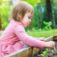 A young child, wearing a pink dress, tends to small plants in a wooden garden bed. The scene is set in a lush, green outdoor environment.
