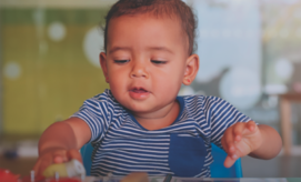 A toddler wearing a striped shirt reaches for toys on a table, in a brightly-lit indoor setting with blurred colorful background elements.