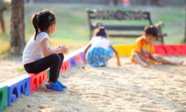A child sits on a colorful sandbox edge, watching two other children play in the sand. A park bench and grassy area are visible in the background.