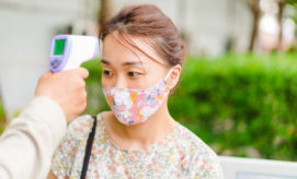 A thermometer measures a masked person's temperature outdoors. The individual is wearing a floral-patterned mask and top. Vibrant greenery and a blurred building provide an outdoor backdrop.