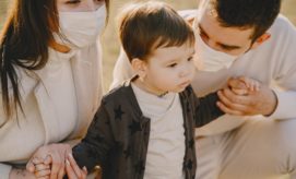 Adults wearing masks hold the hands of a young child standing between them outdoors. The child, in a star-patterned jacket, looks ahead with a neutral expression.
