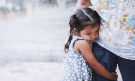 A young girl in a polka-dot dress hugs an adult's leg, standing outdoors on a paved surface with blurred natural surroundings.