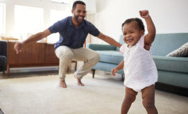 A toddler in a light outfit joyfully raises an arm while walking in a living room. An adult crouches behind, smiling broadly, near a blue sofa and large windows.