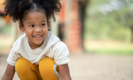 A young child crouches and smiles, wearing a speckled white sweater and yellow pants. The background shows a blurred park setting with trees and structures.