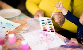 A person holds a paintbrush, guided by a child, over a watercolor palette on a colorful, paint-splattered table. Nearby are pink paint bottles and abstract painted papers.