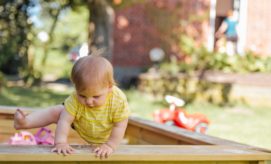 A toddler in a yellow striped shirt balances on a wooden structure, extending a leg upward. In the background, a garden scenery with a blurred red toy car and building.